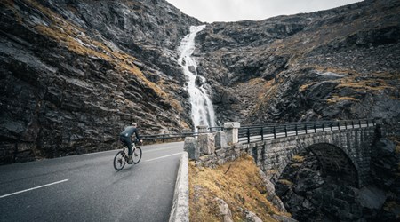 Cyclist riding along a mountain road with a tall waterfall cascading down the cliffside in Sunnmøre, Norway