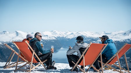 Friends relaxing in deck chairs on a snowy mountaintop, enjoying the fjord view in winter sunshine
