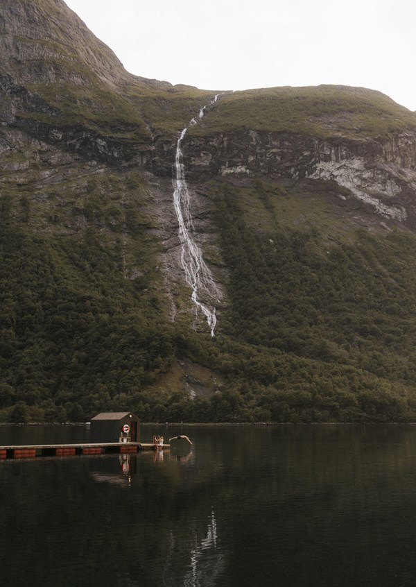 Floating sauna on a tranquil fjord beneath a mountain waterfall